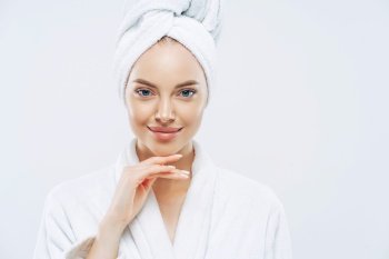 Cropped shot of young good looking European woman looks with calm face, enjoys bath procedures, wears soft white robe, wrapped towel on head, isolated on white background. Morning time, hygiene