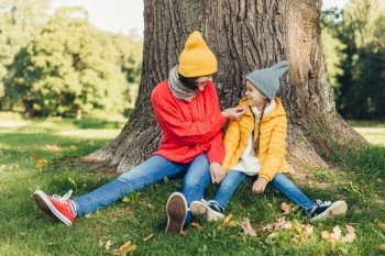Little daughter and her mother have fun together, dressed warm, sit near big tree on green grass, look at each other with love. Affectionate mom and small female kid have good mood, spend weekends
