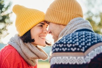 Adorable brunette female and her boyfriend stand closely to each other, look at eyes, smile happily as feel love and support. Good looking woman with scarf and hat has date with young attractive man