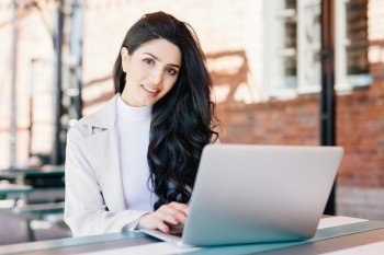 Technology and communication concept. Successful European businesswoman with beautiful appearance working at a cafe on laptop computer having smiling expression looking in camera while sitting outdoor