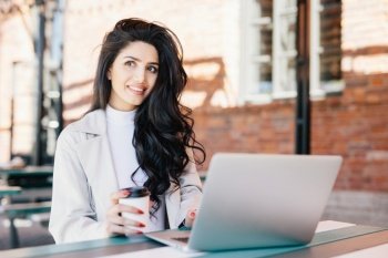 Fashionable brunette woman with long luxurious hair dressed elegantly looking into distance with thoughtful expression dreaming of holidays while working on laptop at outdoor cafe. Technology concept