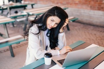 Lovely female businesswoman with dark long hair holding pen in her hand sitting outdoors using laptop computer for her work drinking coffee trying to make project. Adorable brunette female working