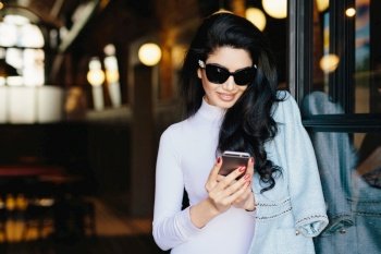 Horizontal portrait of magnificent brunette woman in white clothes and sunglasses sitting in cafe using her smartphone messaging with friends using free internet conncection. Beauty and youth concept