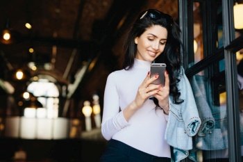 Adorable brunette woman with happy look having sunglasses on head and wearing white blouse siting over modern cafe interior, using cell phone, checking newsfeed on her social network accounts.