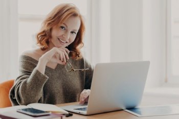 Pleased female scientist makes research on laptop computer, poses in coworking space, searches information for course work, wears warm sweater, prepares for academy test with modern technologies