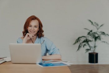 Cheerful foxy female entrepreneur chats online with business partners, works on startup project, poses at workplace, surrounded with paper documents, white wall and potted flower in background