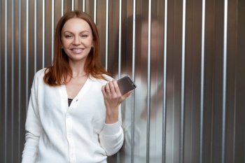 Cute businesswoman waits for call, holds modern cell phone, dressed in white jumper, has brown hair, connected to wireless internet, enjoys advanced technology, receives notification from friend