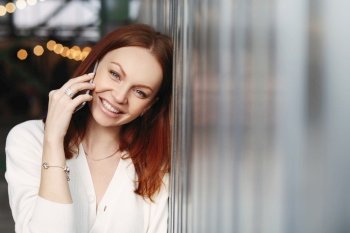 Photo of lovely woman with reddish hair, positive smile, has telephone talk with customer, calls to friend, dressed in white shirt, looks at camera with satisfied expression, enjoys communication