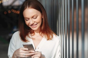 Pleased woman focused into screen of cell phone, checks email box, dressed in white clothes, sends feedback, connected to wireless internet, has brown hair, charming smile, sends text message