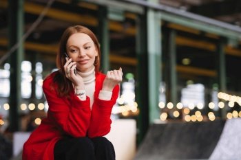 People, technology and lifestyle concept. Positive Caucasian woman has telephone conversation, talks to friends via cellular, wears red coat and black trousers, poses outside with free space