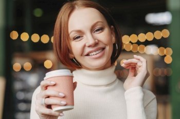Brown haired woman with make up, toothy smile, dressed in white turtle neck sweater, enjoys hot drink, smiles positively, poses in coffee shop, has make up, expresses happiness. Drinking concept