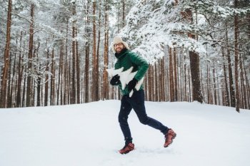 Optimistic young male dressed in warm winter clothes, has fun outdoor in winter forest, breath fresh air, glad to see much snow, holds atrficial fir tree in hands. People, season, weather concept