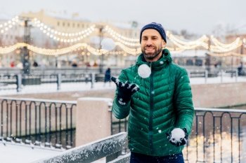 Outdoor shot of happy smiling bearded male in warm jacket and hat plays snowballs as stands against beautiful lights, being in good mood, has joyful expression. People, winter and fun concept