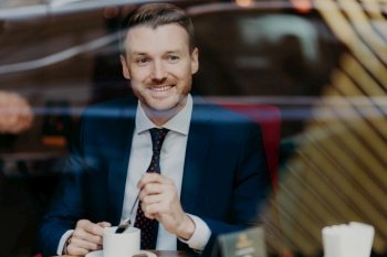 Horizontal shot of happy businessman dressed in formal clothing, drinks aromatic hot beverage, looks at window while sits in cafeteria, has pleased expression. People, rest and lifestyle concept