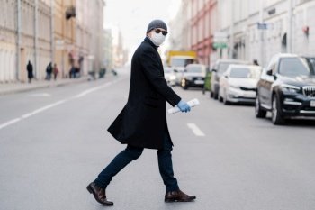 Virus covid -19. Man crosses road, dressed in black coat, hat and sunglasses, wears ptotective mask for pandemic virus protection, poses outdoor at busy street with many transport and people