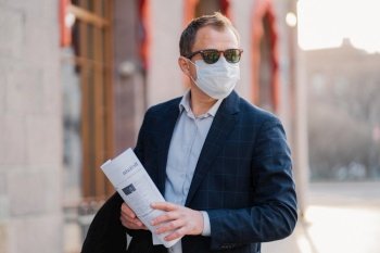 Businessman wears protective mask against transmissible infectious disease, reads newspapers, dressed elegantly, poses outdoor, thinks how to prevent new coronavirus from China. Flu in city.