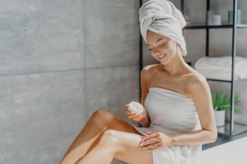 Photo of sensual young European woman uses body cream after taking bath takes care of her skin wrapped in white towel poses against bathroom background. Wellness, pampering and wellbeing concept