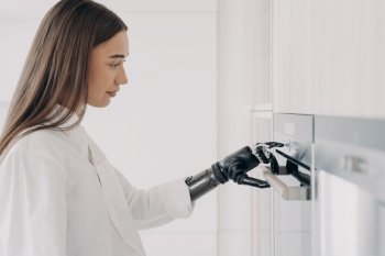Disabled girl presses button on oven by finger of bionic prosthetic arm. Woman with artificial hand cooking preparing dinner with modern kitchen appliance. High tech prosthesis advertising.. Disabled girl presses button on oven by finger of bionic prosthetic arm. High tech prosthesis ad