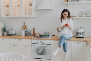 Happy woman housewife sitting in modern clean cozy kitchen at home with smartphone. Smiling female ecommerce client shopping online, enjoy resting after chores, satisfied with house cleaning services.. Female housewife satisfied with house cleaning services sits in modern clean kitchen with smartphone