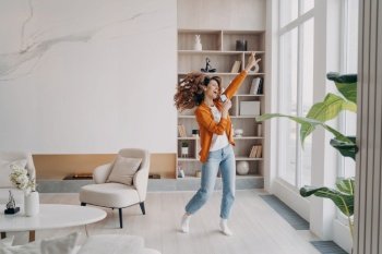 Energetic woman holding smartphone, having fun, dancing in modern living room, excited female dances to music on phone, singing, rejoicing with good news spending leisure time at home.. Energetic woman holding smartphone, having fun, dances to music rejoicing with good news at home