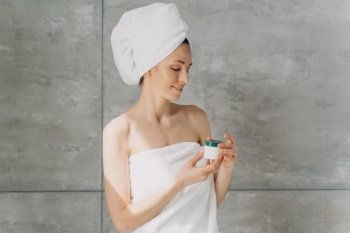 Young woman in towel holds cosmetic jar with cream for healthy facial skin. Female with naked shoulders holding serum in hands in bathroom. Rejuvenation skincare beauty product advertising. Copy space. Young girl in towel holds cosmetic jar with cream in bathroom. Skincare beauty products advertising