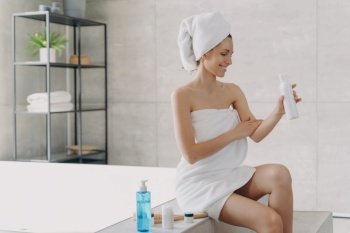 Smiling woman wrapped in towel applying organic body care lotion sitting on bathtub in modern bathroom. Female using sunscreen cream after shower. Self-care, skincare treatment, spa procedure. Smiling girl apply body care lotion sitting in modern bathroom. Skincare treatment cosmetics ad