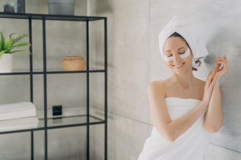 Woman wrapped body and head in towels posing with under eyes patches for skin after shower in bathroom. Female with closed eyes enjoying skincare treatment. Anti-wrinkle hydrogel patch advertising.. Female wrapped in towels with under eyes patches on skin enjoying skincare treatment in bathroom