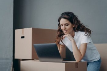Young hispanic girl delivery service worker working at laptop surrounded by cardboard boxes. Businesswoman online store owner preparing customer orders parcels for sending. Small business, ecommerce.. Businesswoman works at laptop prepares online store orders for sending. Small business, ecommerce