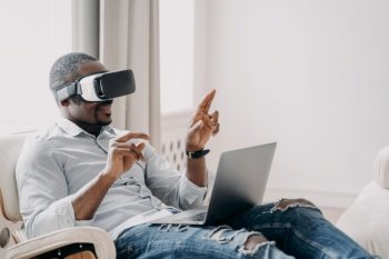 African american businessman wearing VR glasses working at laptop, interacting with virtual reality sitting in armchair. Modern black man touching object in cyberspace. High tech concept.. African american businessman in virtual reality glasses working at laptop, sitting in armchair