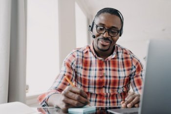 African american male student wearing headset learning foreign language online at laptop sitting at desk. Black man in headphones listening lecture, making notes. E learning, distance education.. African american male student in headset makes notes, learning online at laptop. Distance education