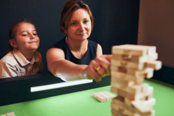 Mother playing jenga game with her daughter in play room. Woman removing block from stack. Unstable tower of wooden blocks. Game of skill and fun. Family time