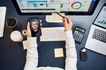 Businesswoman having video chat on smartphone with her client. Businesswoman working with data on charts, graphs and diagrams on computer screen. Woman holding smartphone sitting at desk in office