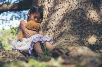 Sad girl hugging teddy bear sitting under tree sadness alone in green park. Lonely girl feeling sad unhappy sit outdoors hug best friend toy. Autism child play teddy bear best friend. Family violence