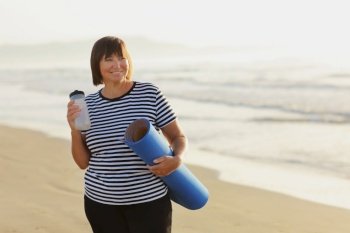 Smiling mature adult woman with bottle of water after exercising outdoors holding yoga mat outdoors on beach by sea on summer. Senior healthy lifestyle, vitality, healthcare concept. copy space.. Smiling mature adult woman with bottle of water after exercising outdoors holding yoga mat outdoors on beach by sea on summer. Senior healthy lifestyle, vitality, healthcare concept. copy space