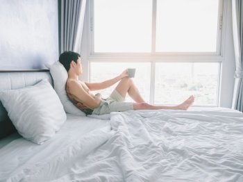 Asian young man with tattoo holding coffee cup on the bed looking out window in morning with sunrise light