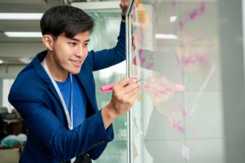 Young Creative businessman holding a marker and writing plan and share idea on glass wall with sticky note, Brainstorming and discussing and formulating, business strategies in tech startup office.