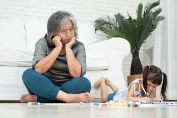 Exhausted old elderly grandmother sit on floor in living room and feel unwell tired from little children running and playing loud, suffer from headache, female nanny feels exhausted by noisy kids