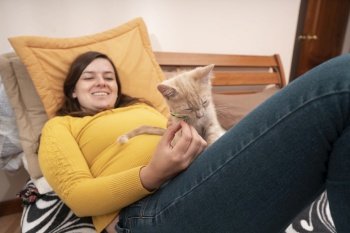 Beautiful Hispanic young woman smiling dressed in yellow jacket, lying on her bed playing with her little kitten. focused cat. Beautiful young Hispanic woman lying on her bed playing with her little kitten