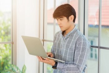Asian businessman smiling and working on a laptop computer stand near window in office. Portrait of handsome man standing and online meeting with computer notebook in office lobby