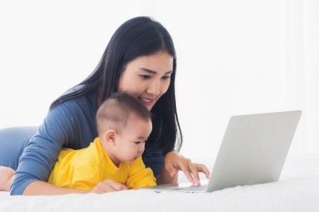 Asian happy young mother and baby lifestyle working on laptop, woman mom and daughter reading and learning technology on screen while notebook computer on bedroom white bed