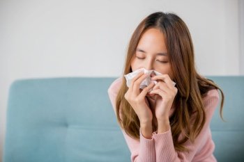 Ill Asian young woman cold covered with blanket sitting on sofa and sneeze with tissue paper at home, Female sick allergic blowing nose sneezing in tissue at home, Flu health care