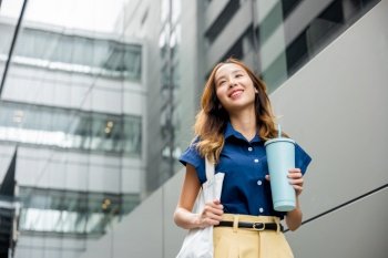 Asian beautiful business woman confident smiling with cloth bag holding steel thermos tumbler mug water glass she walking outdoors on street near modern building office, Happy female looking side away