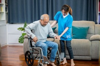Asian nurse assisting helping senior man patient get up from wheelchair for practice walking at home, Smiling doctor support old man to getting up, help handicapped elderly stand up, healthcare