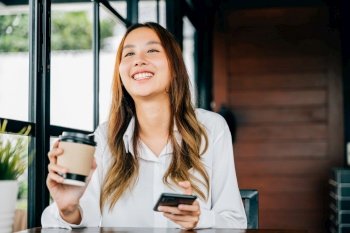 Asian young business woman holding coffee paper cup and using mobile phone to online chating at coffee shop, Happy female sitting in cafe near window she holding mobile phone on hand