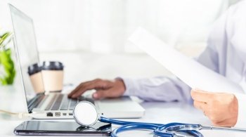 Closeup of doctor or nurse woman in uniform with stethoscope typing information of patient prescription from paperwork medical history form into laptop computer for record document data report