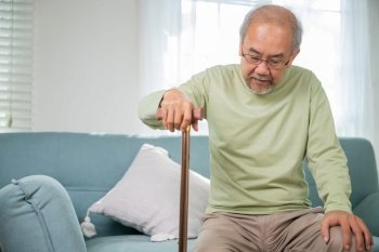 Asian Old man with eyeglasses typing to stand up from sofa with walking cane, Elderly suffering from knee pain ache holding handle of cane, senior disabled man holding walking stick at home