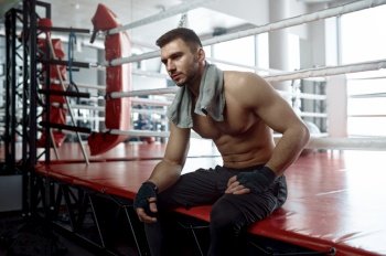 Portrait of muscular male boxer sitting on boxing ring mat and rest after hard training session or competitive match. Muscular male boxer sitting on boxing ring mat