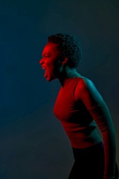 Headshot portrait of emotional young woman crazy shouting holding head with hand in neon light over dark studio background. Headshot portrait of young woman shouting