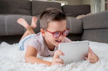 Cute little boy watch cartoons on digital tablet. Kid lies on floor laughing using electronic device. Indoor leisure for children. High quality photography. Cute little boy watch cartoons on digital tablet. Kid lies on floor laughing using electronic device. Indoor leisure for children.
