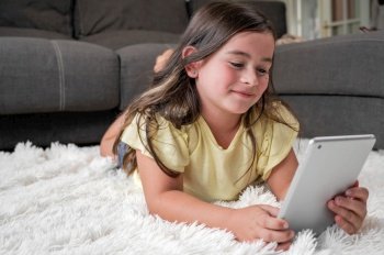 Cute little girl watch cartoons on digital tablet. Kid lies on floor laughing using electronic device. Indoor leisure for children. High quality photography.. Cute little girl watch cartoons on digital tablet. Kid lies on floor laughing using electronic device. Indoor leisure for children.